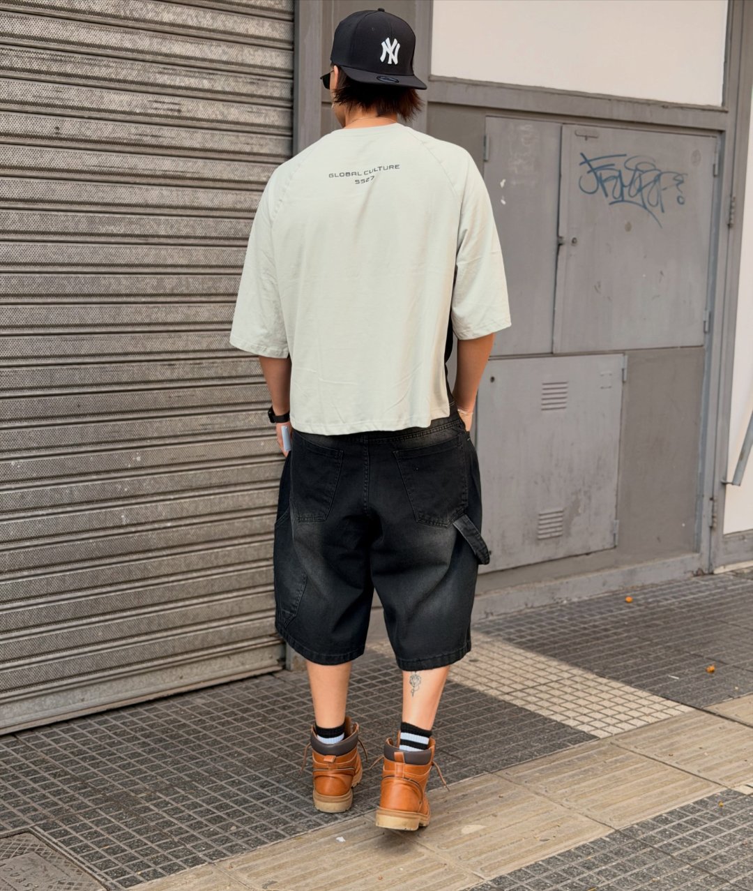 Espalda de la remera gris con la estampa Global Culture SS27, gorra negra y bermuda de jean